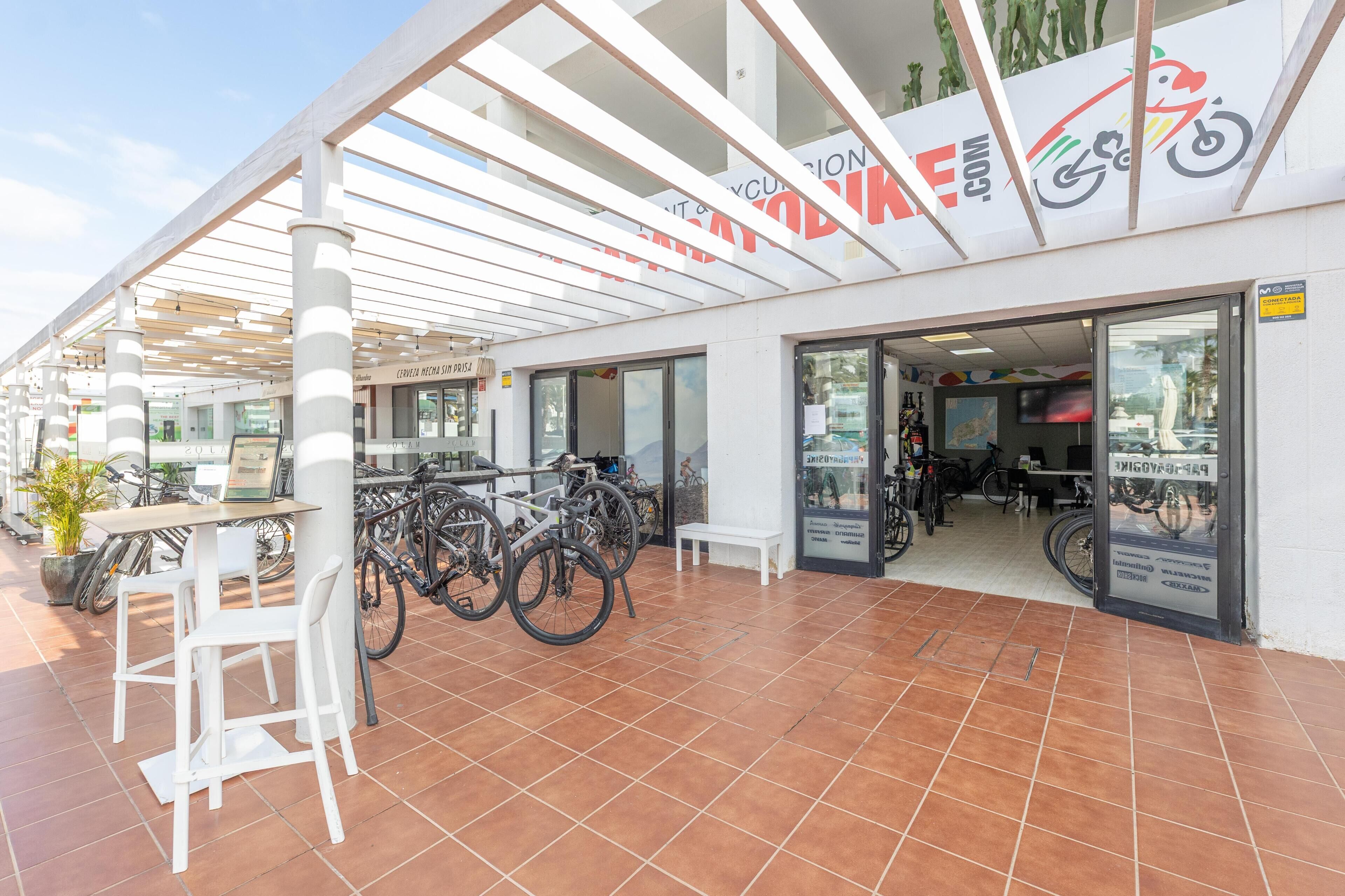 A covered outdoor area with a bike rental service, a seating area with white chairs, and a tiled floor.