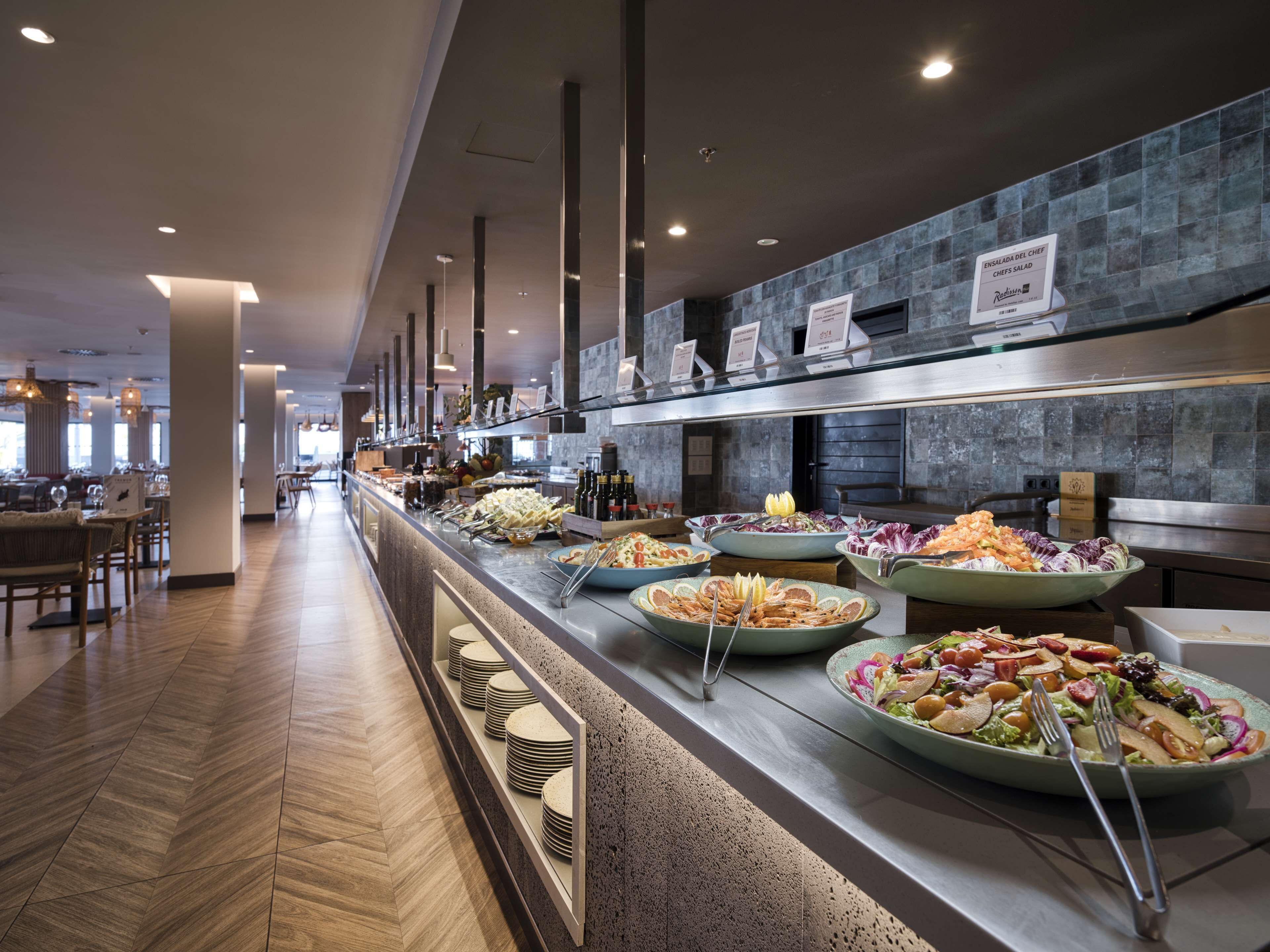 Un espace buffet où diverses préparations sont présentées sur un comptoir, un coin repas avec des tables et des chaises, et un aménagement intérieur moderne avec un motif de parquet en chevrons.