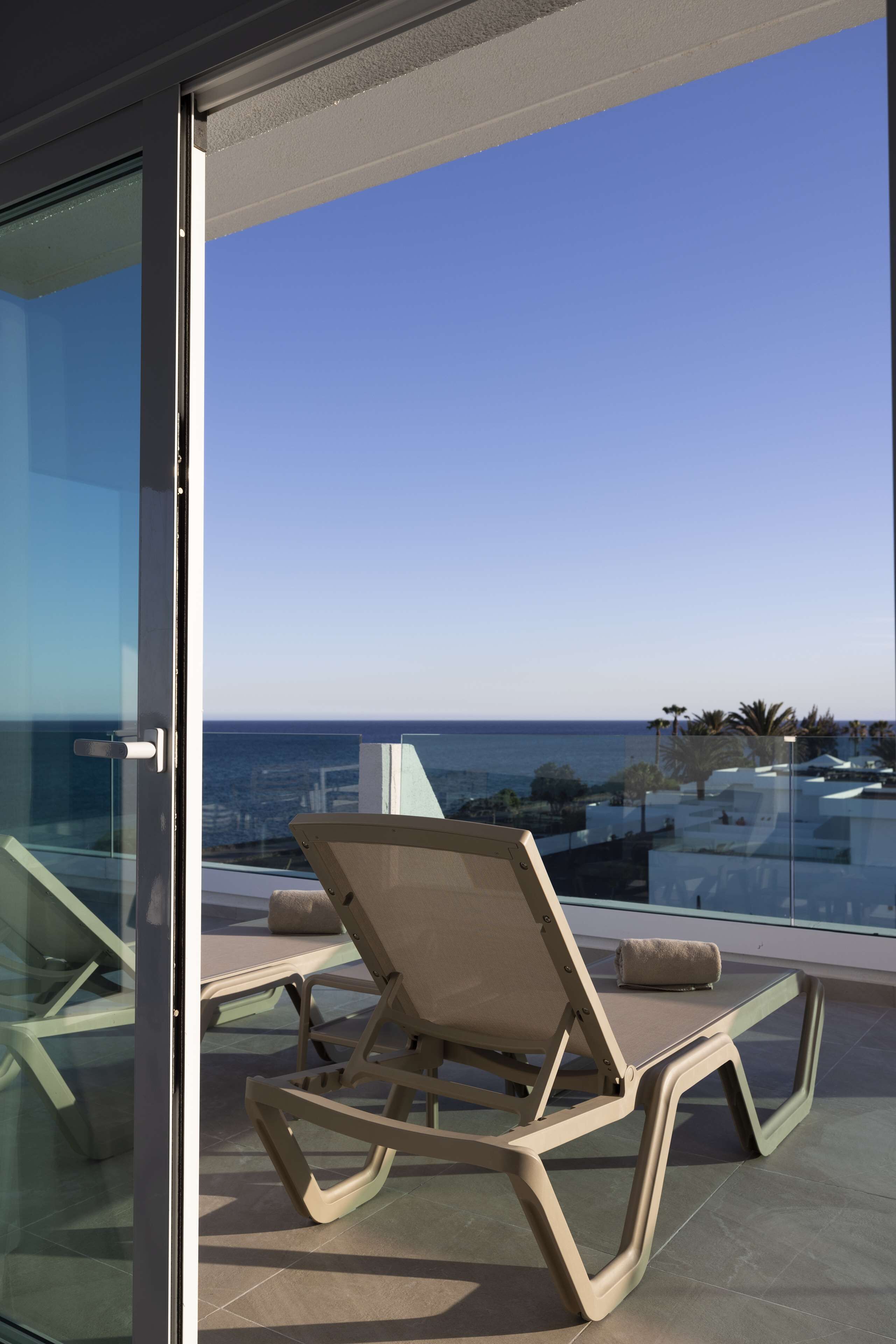 A balcony with a glass door, a lounge chair, and a view of the ocean and cityscape.