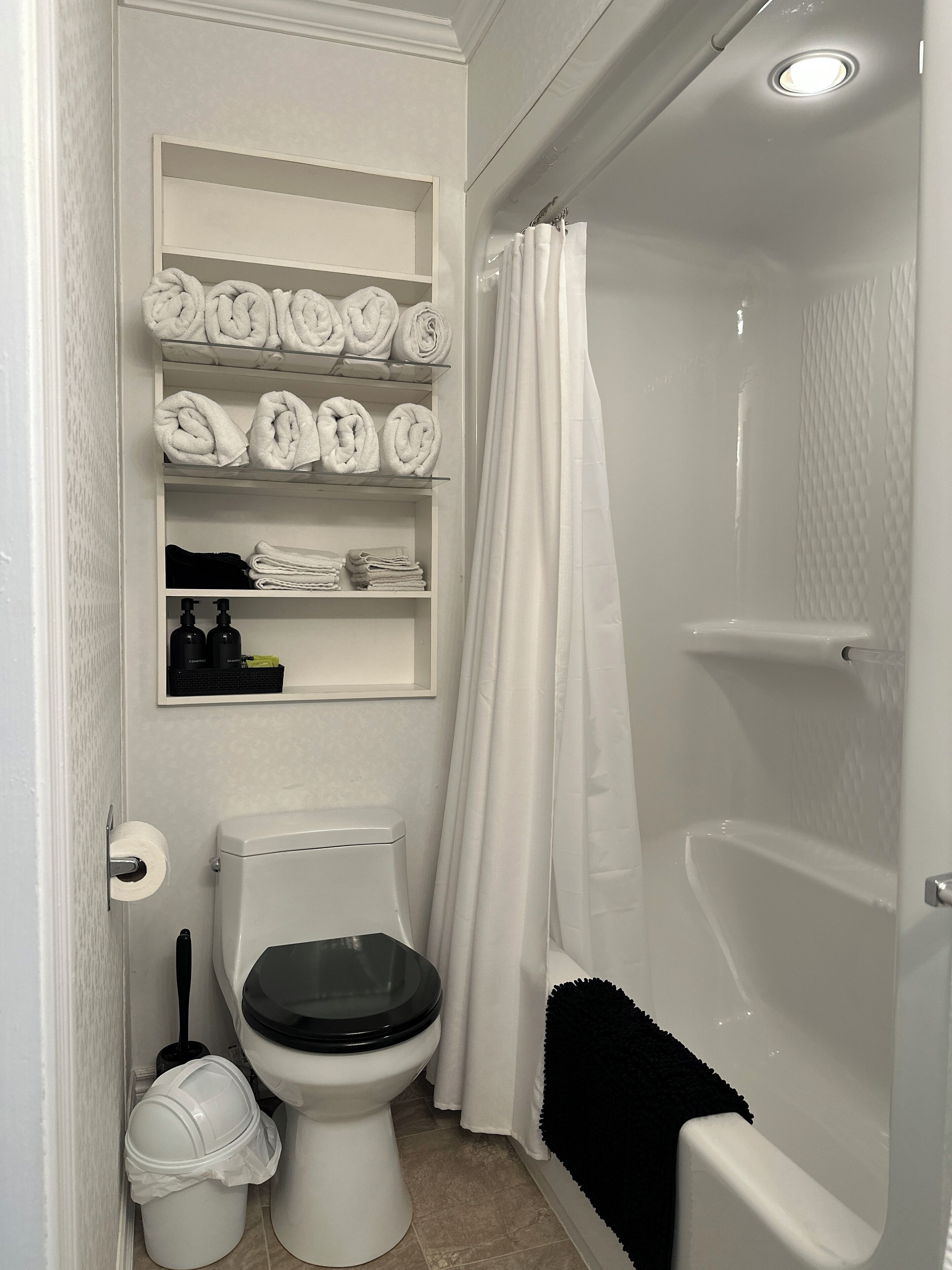 Une salle de bain avec des toilettes, une étagère où sont pliées soigneusement des serviettes, un rideau de douche et une brosse pour les toilettes.