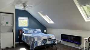 A bedroom with a sloped ceiling, a ceiling fan, a bed with a patterned bedspread, a TV, a heater, and a window with blinds.