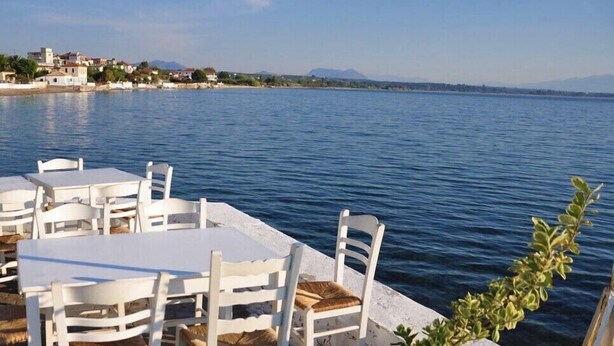Terrassa al mar amb taules i caires blanques, amb vista a un mar tranquil i una costa llunyana amb edificis.