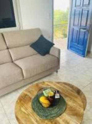 Una sala de estar con un sofá beige, una mesa redonda de madera con frutas, y una puerta azul al fondo.