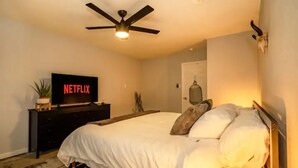 A bedroom with a bed, a ceiling fan, a TV on a stand, a nightstand, and a wall-mounted light.