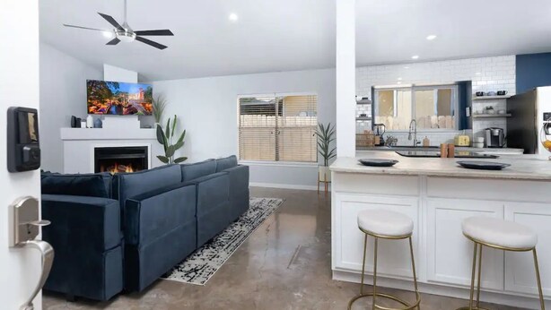A modern living room with a dark blue sofa, a fireplace, and a TV. There's a kitchen with white cabinets and a bar area with stools.