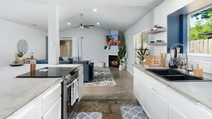 A modern kitchen with white cabinetry, a black countertop, and a built-in oven. There is a dining area with a blue sofa and a rug. A TV is mounted on the wall above the fireplace.