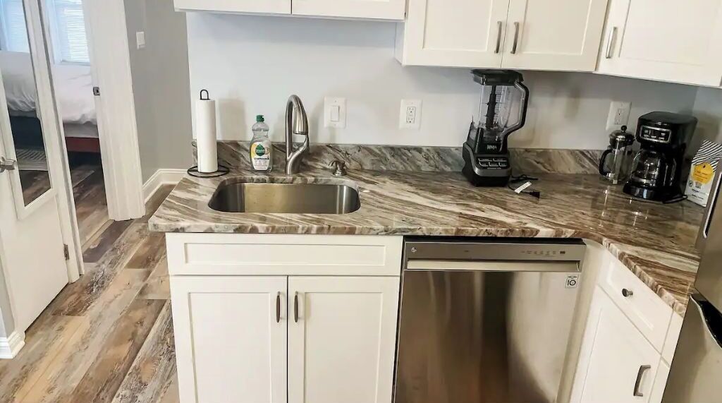 A kitchen with white cabinets, a marble countertop, a stainless steel sink, a dishwasher, a coffee maker, and a blender.