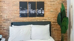 Una cama con almohadas blancas, una pared de ladrillo y tres cuadros de un paisaje urbano blanco y negro encima.