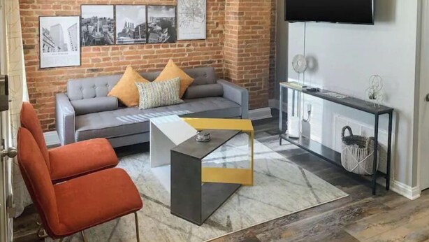 Una sala de estar con pared de ladrillo, un sofá gris con cojines naranjas, un sillón rojo, una mesa de centro y un televisor en la pared.