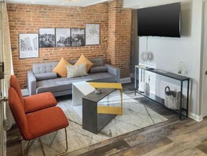 A living room with a brick wall, a grey sofa with orange cushions, a red chair, a coffee table, and a TV on the wall. - Cozy Modern Micro Apt in the Heart of Fells Point! (Baltimore)