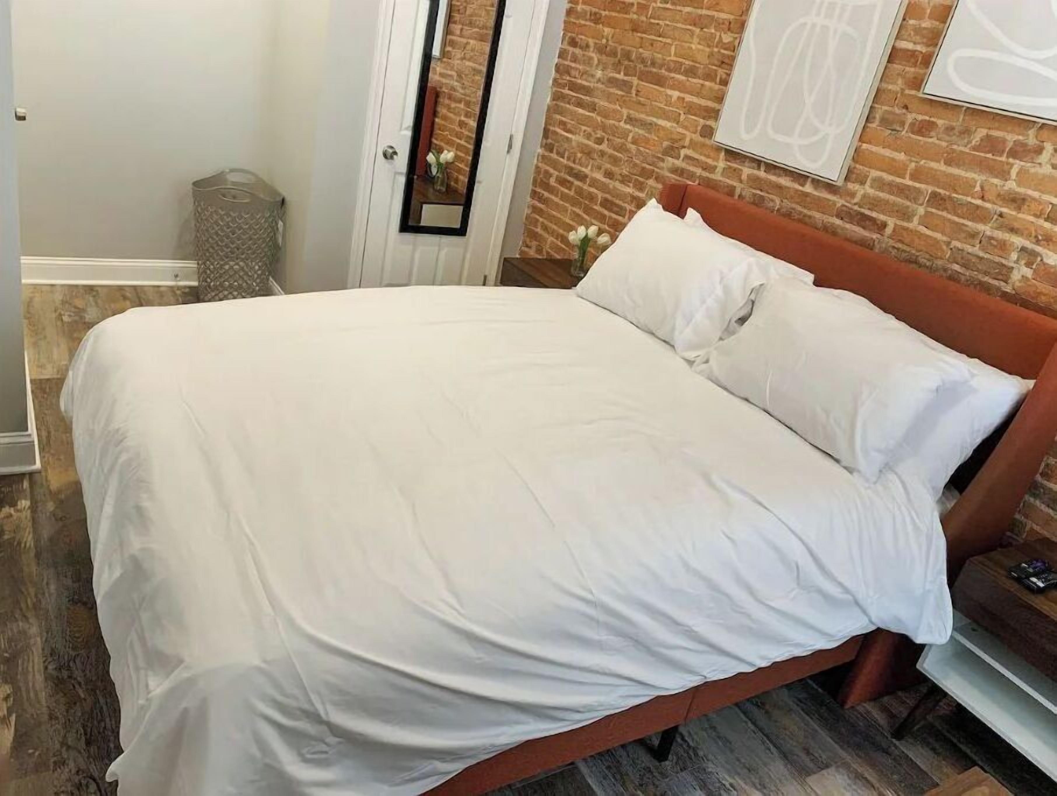 Una cama bien hecha con sábanas blancas en una habitación con pared de ladrillo y suelo de madera.
