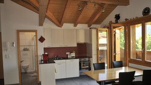 A modern kitchen with white cabinets, a black countertop, and a wooden ceiling. There is a dining area with a table and chairs, and a view through the glass door. - Am Liimerweg by Interhome (Adelboden)