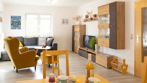 A modern living room with a sofa, a dining table with food and drinks, a TV, and wooden cabinets. - Vacation home Enikö by Interhome (Ostseebad Lubmin)