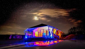 Een grote, verlichte constructie met een reflecterend wateroppervlak, voor een sterrenhemel.