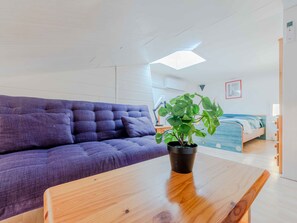 Ein modernes Zimmer mit einem violetten Sofa, einem Holztisch, einer Topfpflanze, einem Bett mit blau-weiß gestreifter Bettdecke und einer weißen Decke mit Oberlicht.