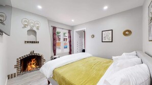 Un dormitorio con chimenea, una cama con ropa de cama blanca y amarilla, una ventana con cortinas y una puerta que da a otra habitación.