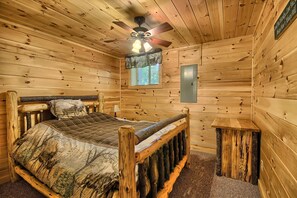 A log cabin room with a bed, a ceiling fan, a window, and a wall-mounted electrical outlet.