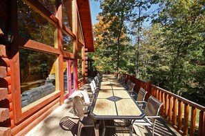A wooden deck with a long table and chairs, surrounded by a wooden railing, overlooking a forested area.