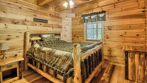 Camera da letto in una baita di legno, con un letto, un comodino, una ventola a soffitto e una finestra con tende.