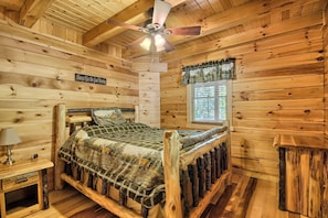 Un dormitorio de cabaña de madera con cama, mesita de noche, ventilador de techo y ventana con cortinas.