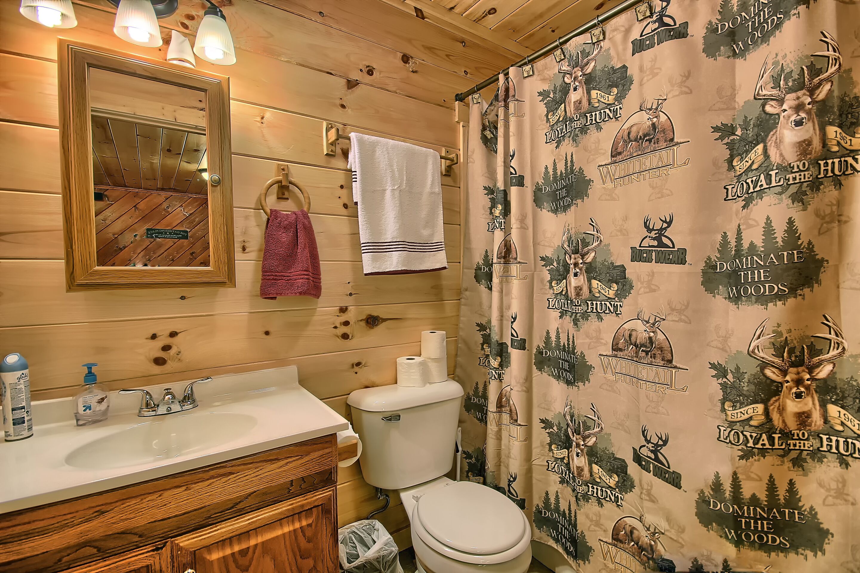A bathroom with wooden walls, a mirror, a sink with toiletries, a toilet, and a shower curtain with hunting-themed designs.