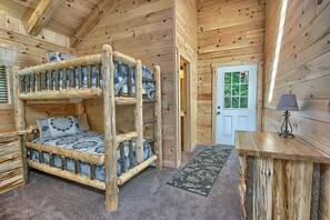 Interior de uma cabana de madeira com uma cama beliche, uma porta, um abajur e uma pequena mesa.