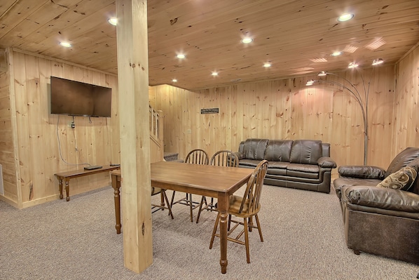 A wooden-paneled room with a dining table, four chairs, two leather sofas, and a flat-screen TV mounted on the wall.