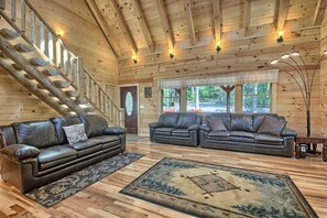 A log cabin interior with two leather sofas, a wooden staircase, and a large window with a view of trees. - Maple Ridge Lodge - 5-Bedroom Lodge -Hocking Hills (South Bloomingville)