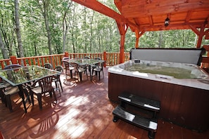 A wooden deck with a hot tub, tables, and chairs, surrounded by trees. - Maple Ridge Lodge - 5-Bedroom Lodge -Hocking Hills (South Bloomingville)