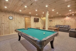 Une table de billard dans une pièce aux murs et au plafond en bois, avec une horloge et un téléviseur.
