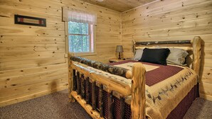 A wooden cabin bedroom with a bed, a nightstand, a lamp, and a window with a view of trees. - Maple Ridge Lodge - 5-Bedroom Lodge -Hocking Hills (South Bloomingville)