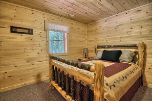 A wooden cabin bedroom with a bed, a nightstand, a lamp, and a window with a view of trees. - Maple Ridge Lodge - 5-Bedroom Lodge -Hocking Hills (South Bloomingville)