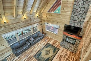 Interior de uma cabana de tronco com lareira de pedra, sofá de couro e uma televisão.