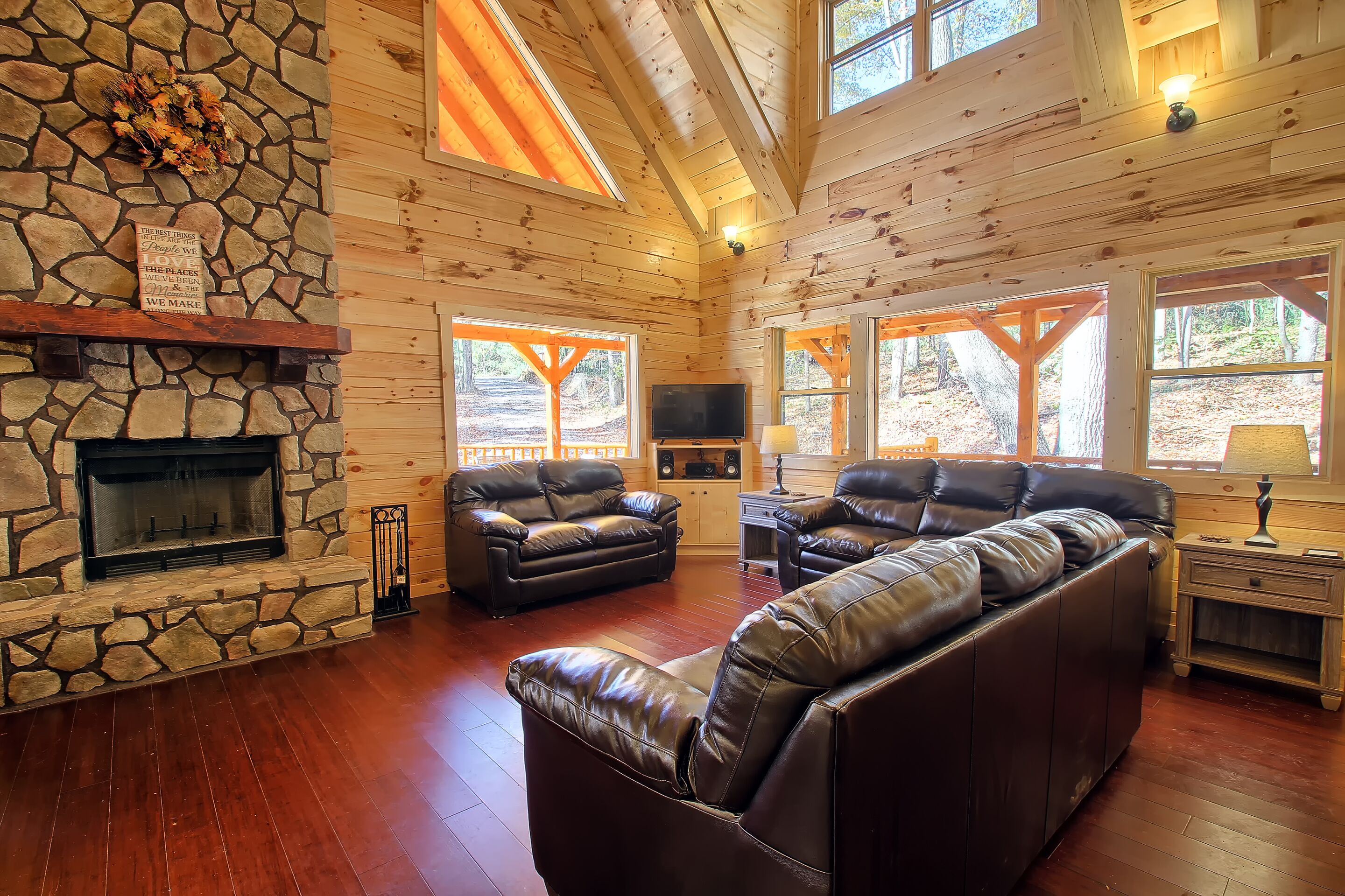 A log cabin interior with a stone fireplace, leather sofas, and large windows.