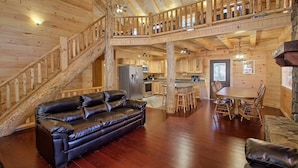 A wooden cabin interior with a leather sofa, a dining table with chairs, a kitchen area, and a staircase leading to an upper level. - Dogwood Lane Lodge- sleeps up to 18 -Hocking Hills (Logan)