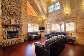 Interior acogedor de una cabaña con chimenea de piedra, sofás de cuero y amplias ventanas con vistas al exterior.