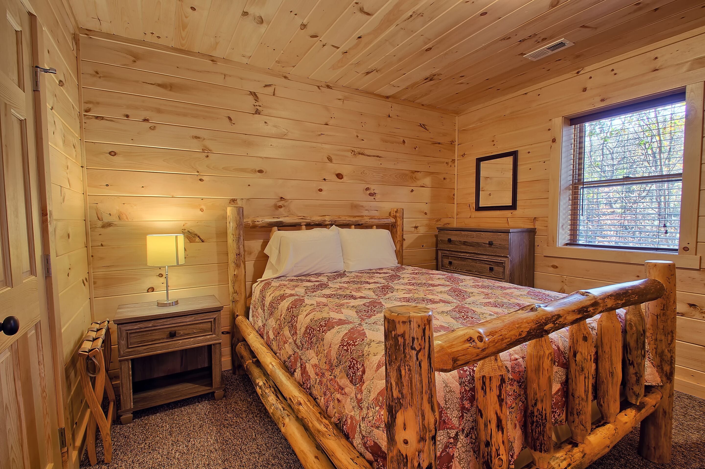 Une chambre à coucher dans un chalet en bois, avec un lit, des tables de chevet, une petite table, une fenêtre avec des stores et un tableau encadré au mur.