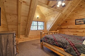 Un dormitorio de cabaña de madera con techo inclinado, una cama con edredón escocés, un aparador y una ventana con vistas a árboles.