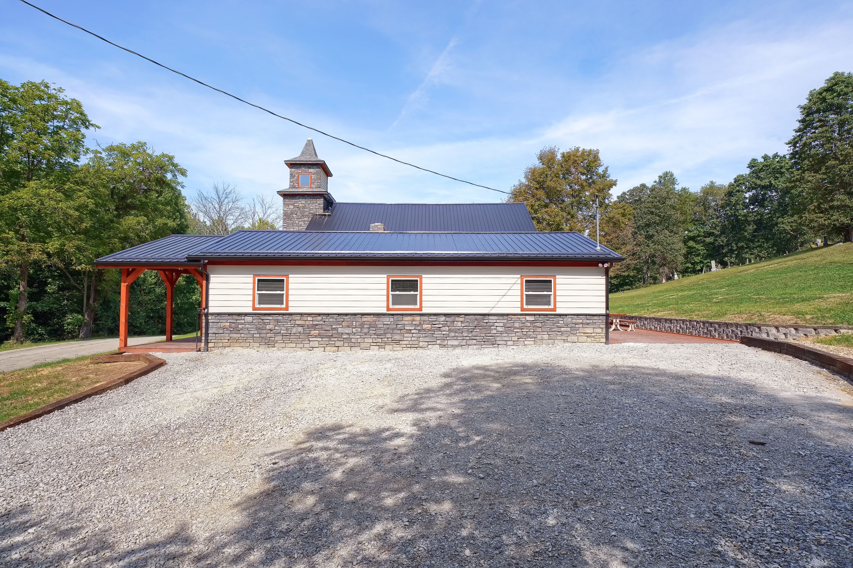 A two-story building with a stone facade and a metal roof, featuring a covered porch and a gravel driveway.
