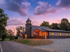 A stone building with a tower and a covered porch, surrounded by trees and a gravel driveway.