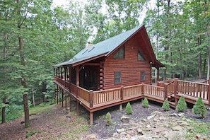 A log cabin with a deck and a stone chimney, surrounded by a forest.