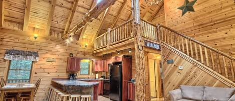 A log cabin interior with a kitchen, dining area, and a staircase leading to a loft.