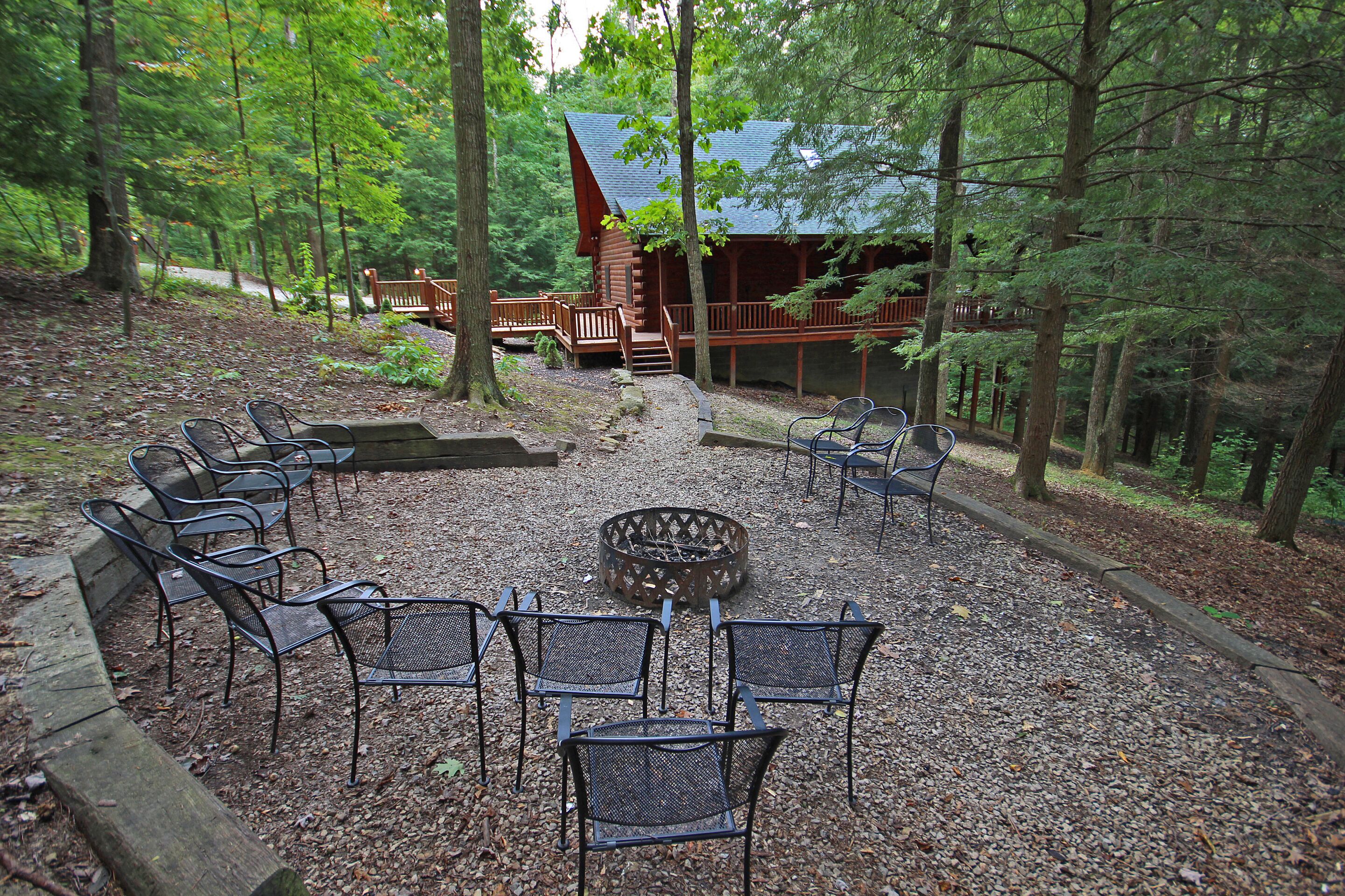 A forested area with a wooden cabin, a fire pit, and metal chairs arranged in a semi-circle.