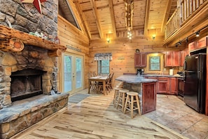 A log cabin interior with a stone fireplace, wooden dining table, and kitchen area.