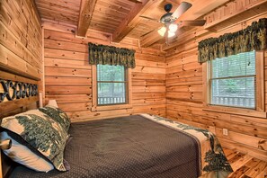 Camera da letto in una baita di legno con un letto, due finestre con tende, una ventola a soffitto e un riscaldatore a parete.
