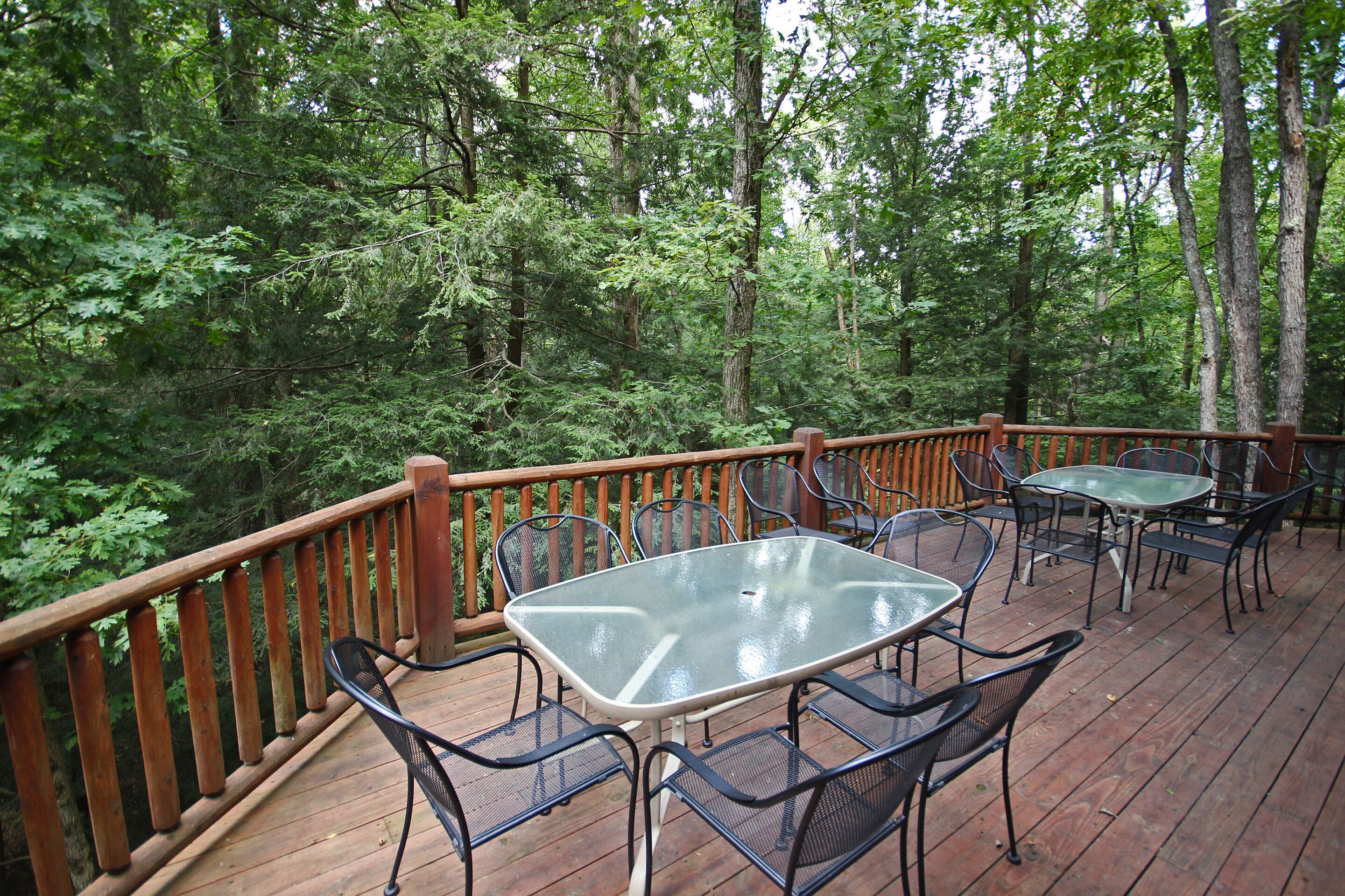 An outdoor deck with a glass-top table and metal chairs, surrounded by a wooden railing, set in a forested area.