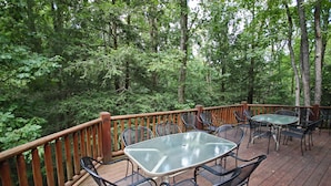 An outdoor deck with a glass-top table and metal chairs, surrounded by a wooden railing, set in a forested area. - Double Pine Lodge - 5 Bedroom -Hocking Hills (Logan)