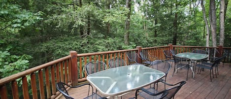 An outdoor deck with a glass-top table and metal chairs, surrounded by a wooden railing, set in a forested area.