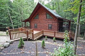 A log cabin with a wooden deck and a porch, surrounded by trees.