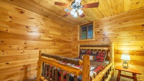 Une chambre dans un chalet en rondins, avec un lit en bois, un ventilateur de plafond, une fenêtre avec des stores et une table de chevet avec une lampe.